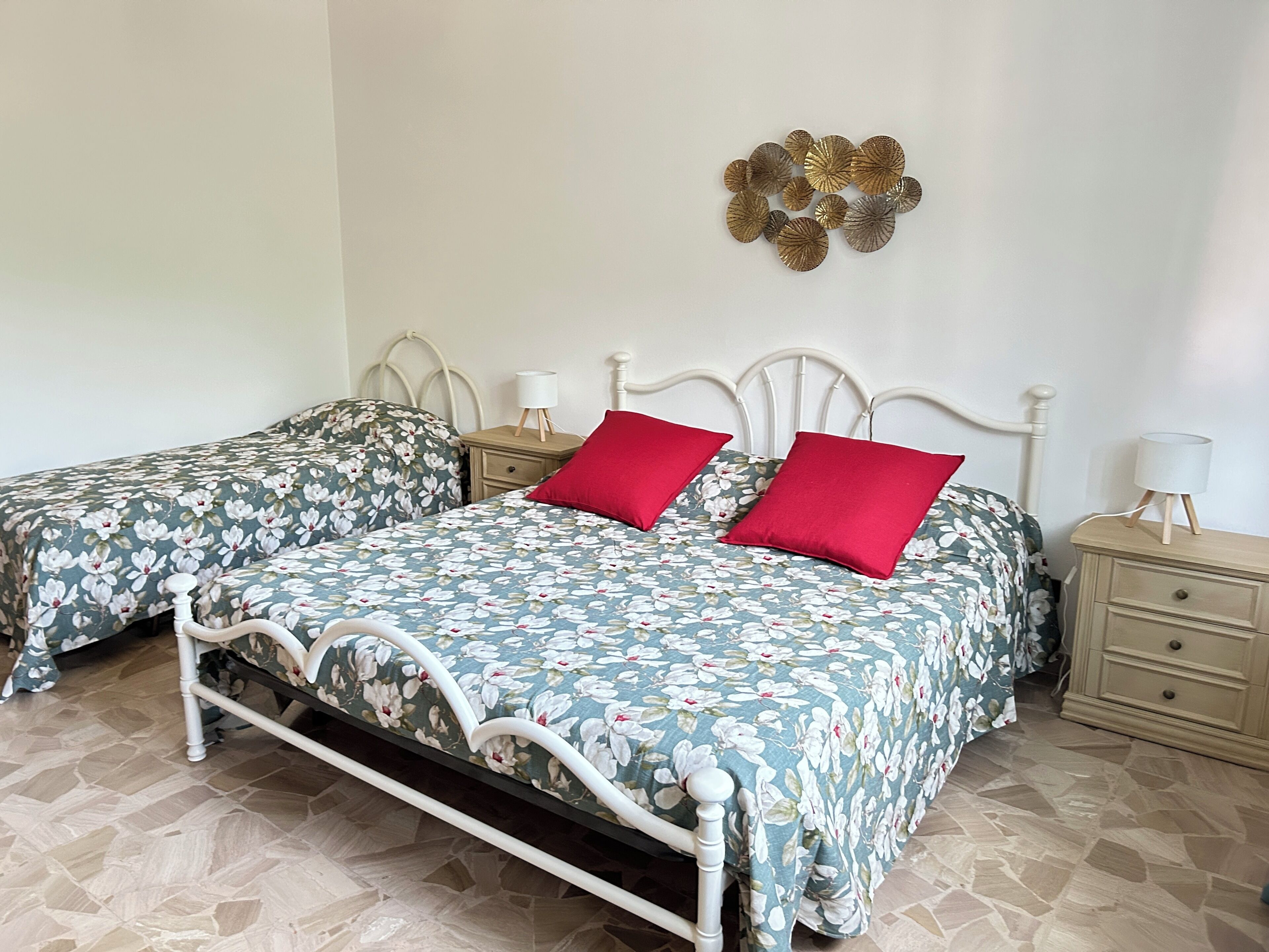 Un lit double avec une literie à motifs floraux et deux oreillers rouges, une table de chevet avec une lampe et un élément décoratif mural.