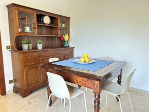 Ein Esstisch mit blauer Tischmatte, umgeben von weißen Stühlen, und ein Holzschrank mit Glastüren und einer Uhr.