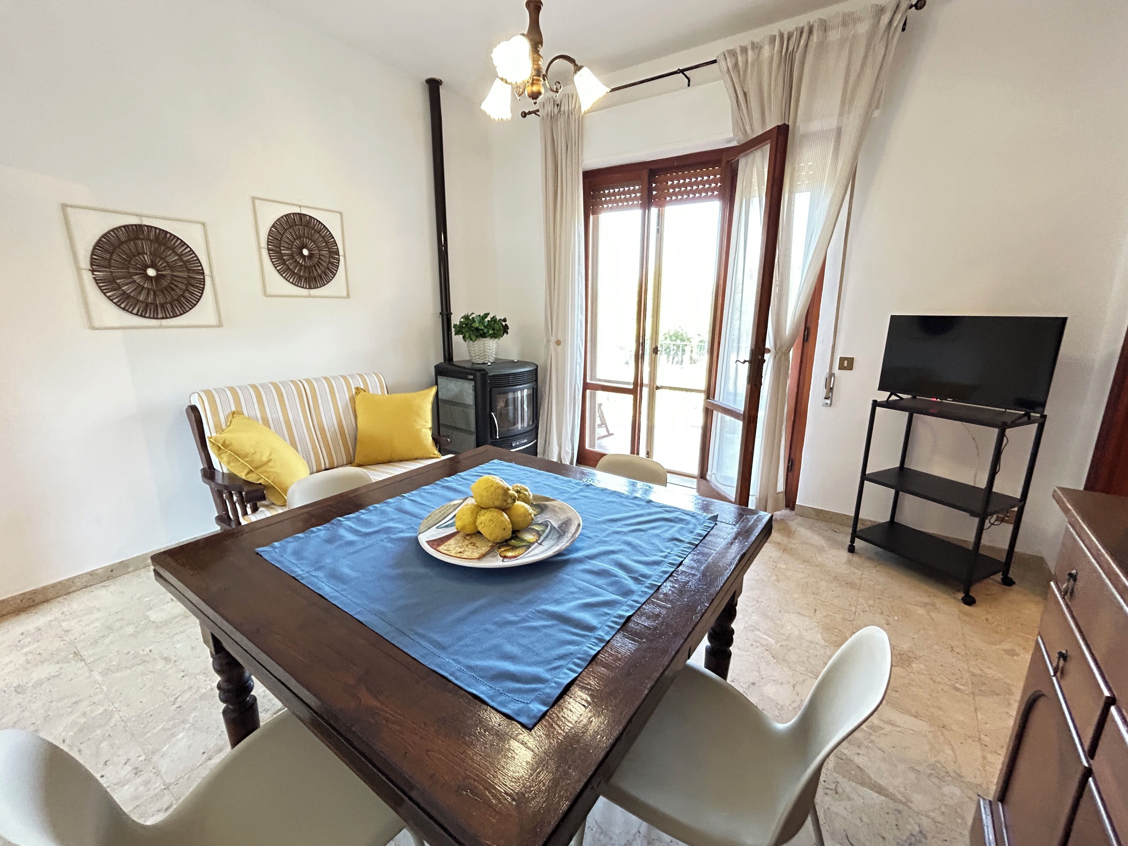 Un coin repas avec une table dressée pour deux personnes, un canapé avec des coussins jaunes, un téléviseur posé sur un meuble et une plante décorative.