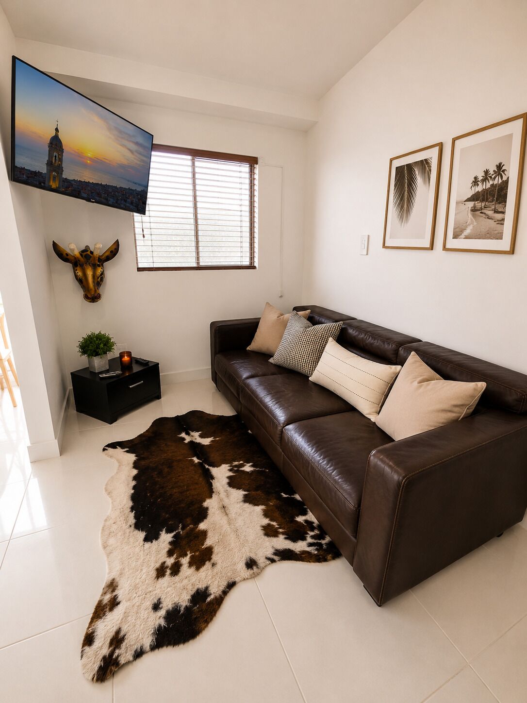 A modern living room with a leather sofa, a cowhide rug, a mounted giraffe head, a TV, and framed pictures on the wall.