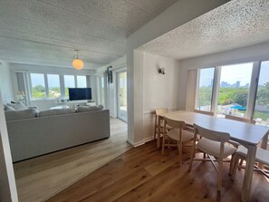Un saló modern amb un sofà, una taula de dinar amb caires, un televisió de pantalla plana i una finestra gran que ofereix vistes a la vegetació i edificis.