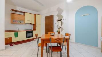 Una zona de menjador amb taula i caires de fusta, una cuina amb armaris de fusta i una porta blava.