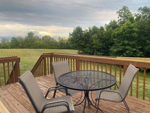 A deck with a round table and four chairs, overlooking a grassy field and trees.