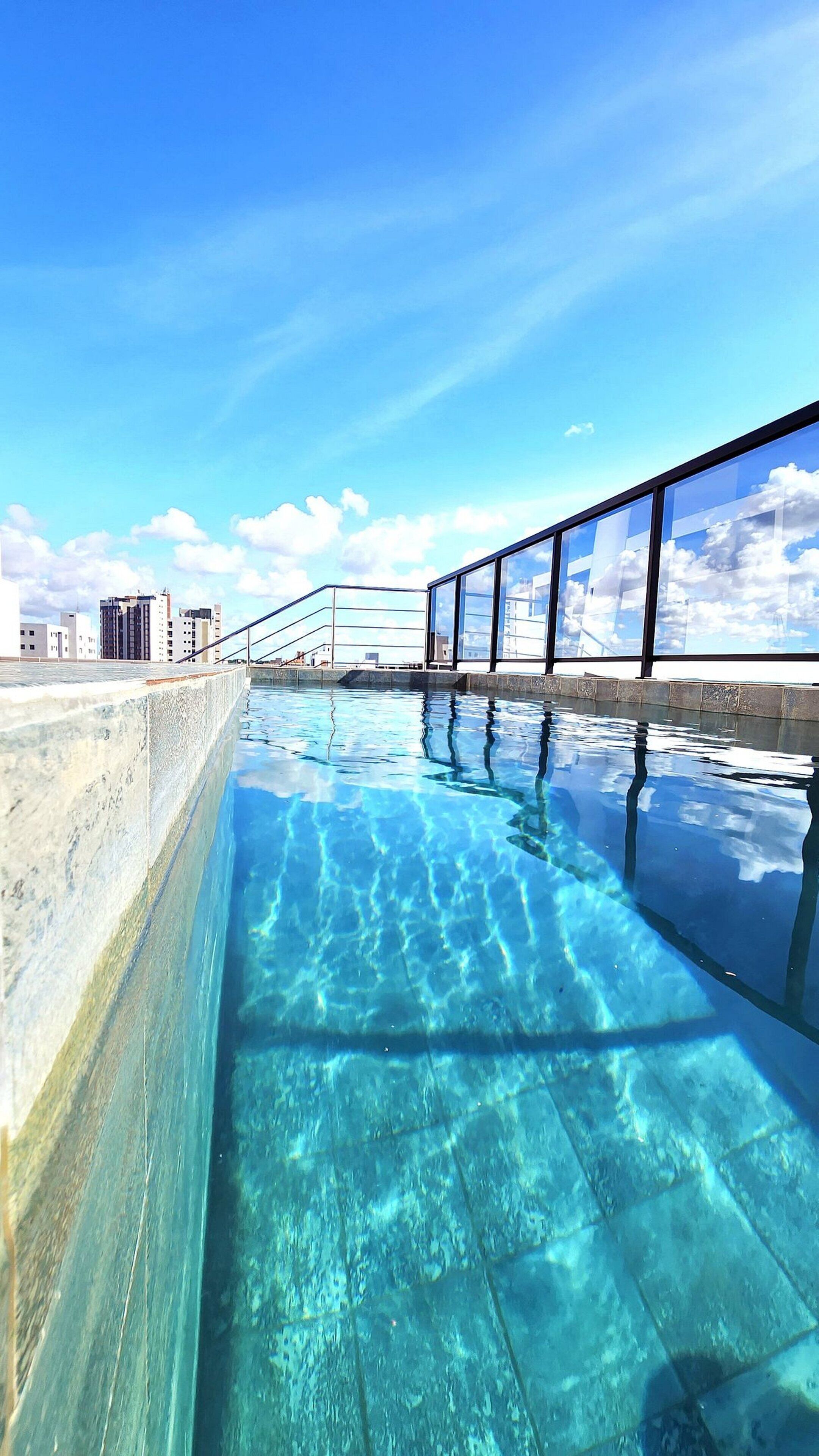 A clear blue swimming pool with a glass barrier and a cityscape in the background.