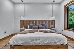 Ein modernes Schlafzimmer mit einem großen Bett, einem Holzkopfteil, zwei Nachttischen und einem Fenster mit Blick auf Grünflächen.