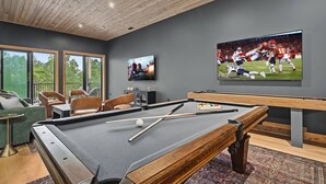 A pool table with cues and balls, a television displaying a sports game, and a seating area with chairs and a sofa. - The Grotto • Indoor Pool • Rooftop • Sleeps 16 (Sevierville)
