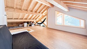 A spacious room with a high ceiling and exposed wooden beams, a hammock, a large window with a view of trees, and a grey sofa.