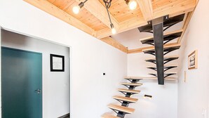 Un escalier en colimaçon avec des marches en bois et des supports métalliques, menant à un plafond en bois avec des poutres apparentes et des luminaires suspendus.