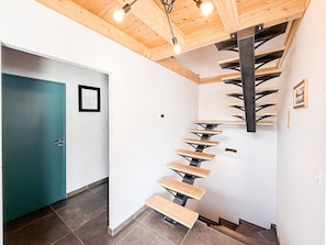 A spiral staircase with wooden steps and metal supports, leading to a wooden ceiling with exposed beams and hanging lights.