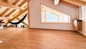 A spacious attic room with wooden beams, a hammock, and a large window.