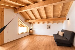 A room with a sloped wooden ceiling, a hammock, a sofa, a small table, and a window.