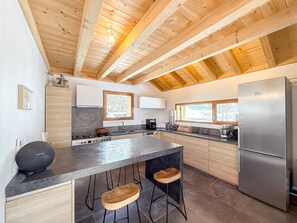 A modern kitchen with wooden beams, a central island, bar stools, and a stainless steel refrigerator.
