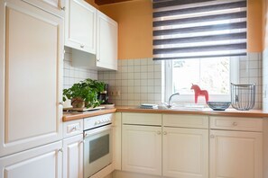 A kitchen with white cabinets, a stove, a sink, and a window with striped blinds. - Ferienhaus 'Ferienhof Homann Sauerland – LH 1' mit Gemeinschaftsterrasse und WLAN (Brilon)