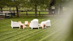 Un espacio al aire libre con césped, sillas de plástico blanco y una mesa pequeña, rodeado de una valla de madera y árboles.