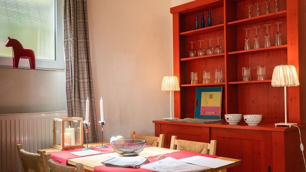 Ein Essbereich mit Holztisch, rotem Tischläufer und Korbstühlen. Ein rotes Bücherregal mit Gläsern ist sichtbar.
