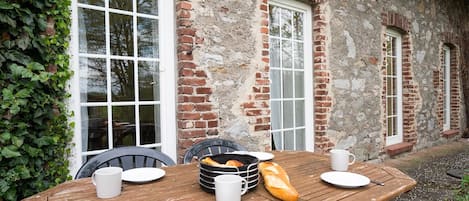An outdoor wooden table with plates, cups, a bread basket, and a baguette.