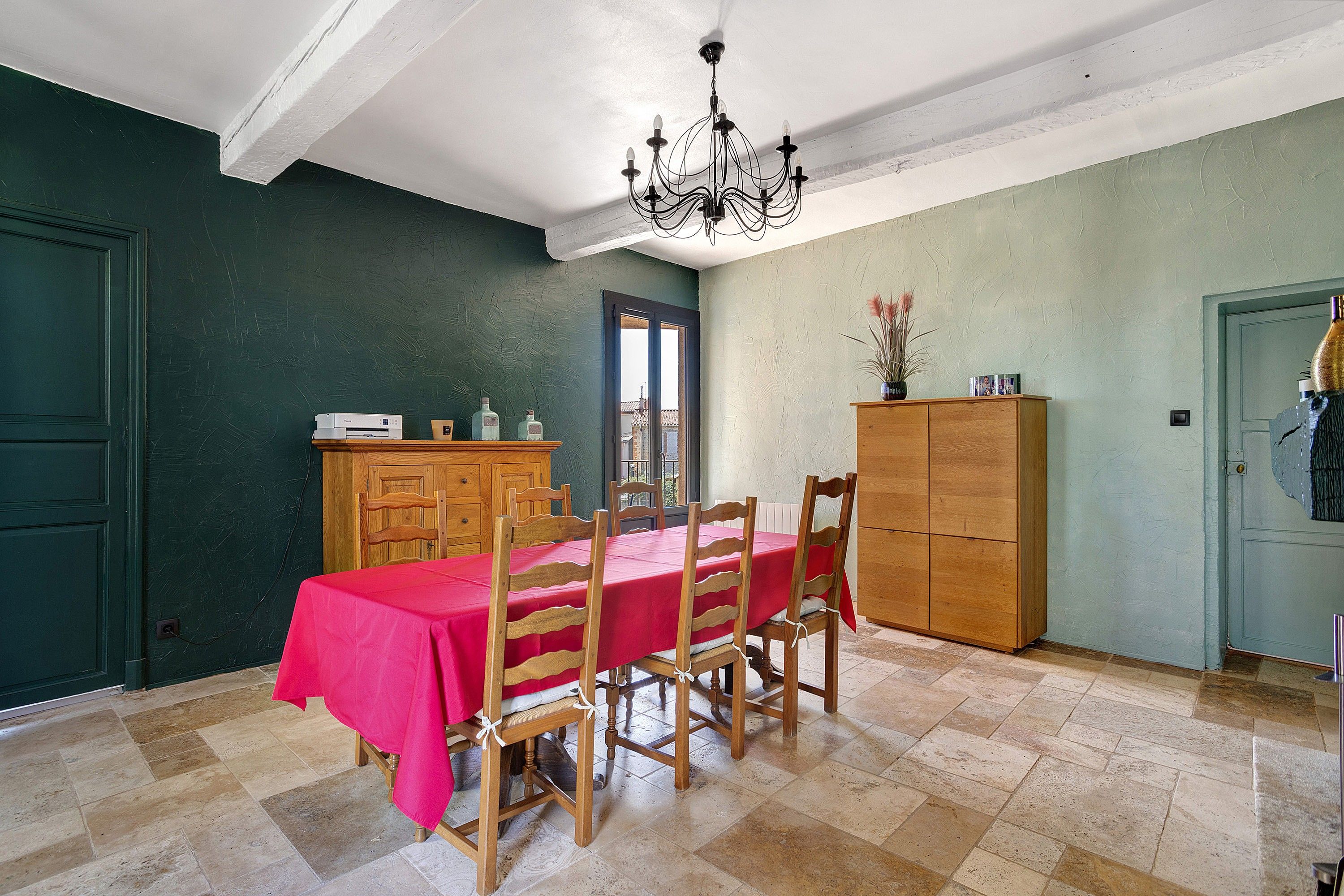 A dining area with a red tablecloth, wooden chairs, a chandelier, a wooden cabinet, and a door with a window.