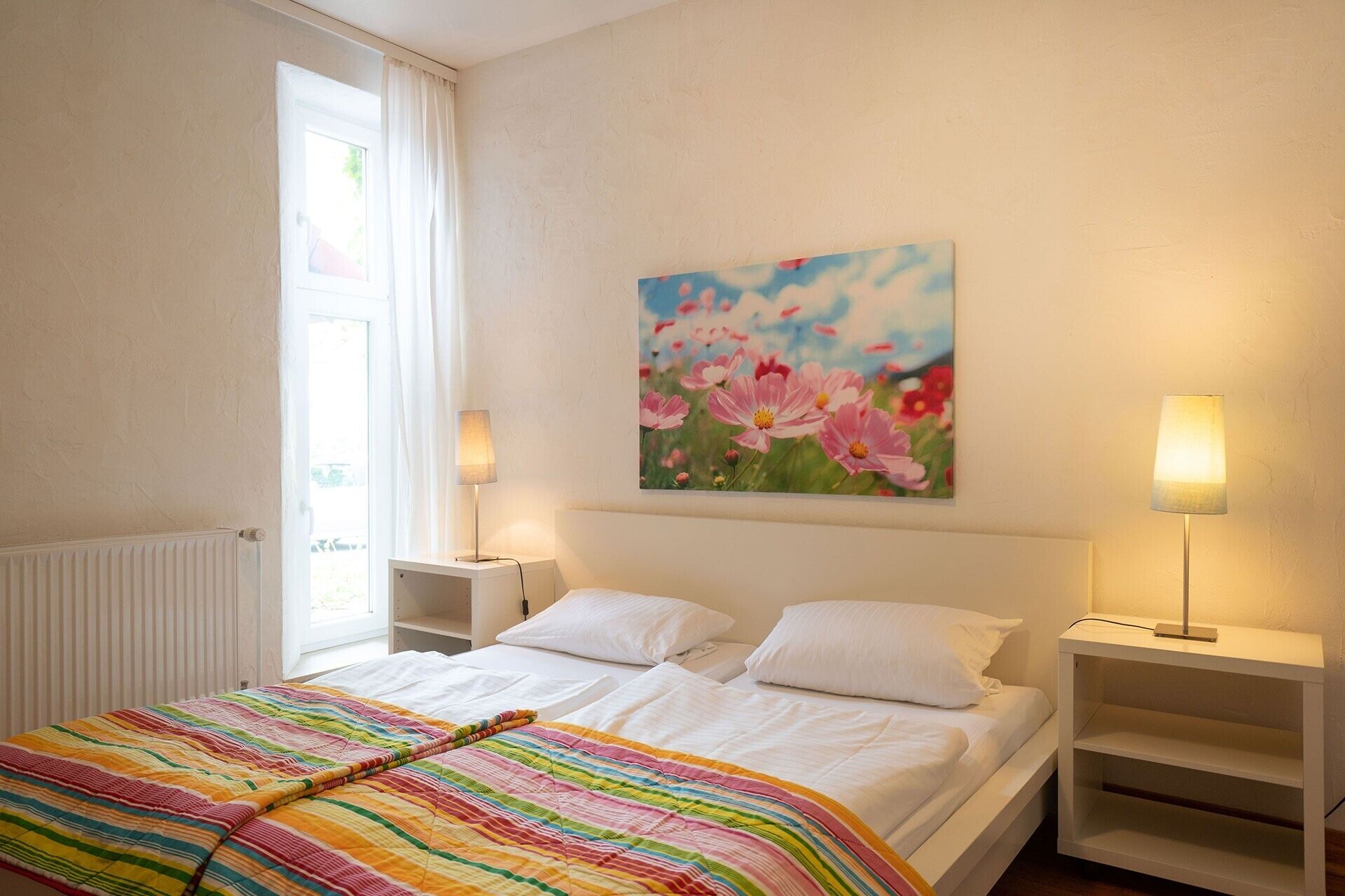 Ein Hotelzimmer mit einem Bett, zwei Nachttischen, einer Heizung, einem Fenster mit Vorhängen und einem Blumenbild an der Wand.