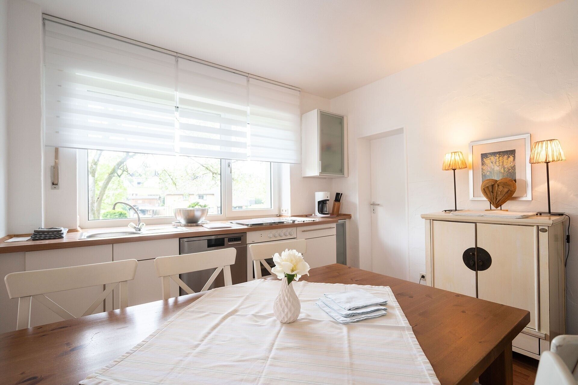 Una cocina moderna con comedor, que incluye una mesa de madera, sillas blancas y un jarrón con flores.