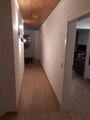 Un pasillo con suelo de madera, paredes blancas y una puerta que da a una habitación con cama y televisor.