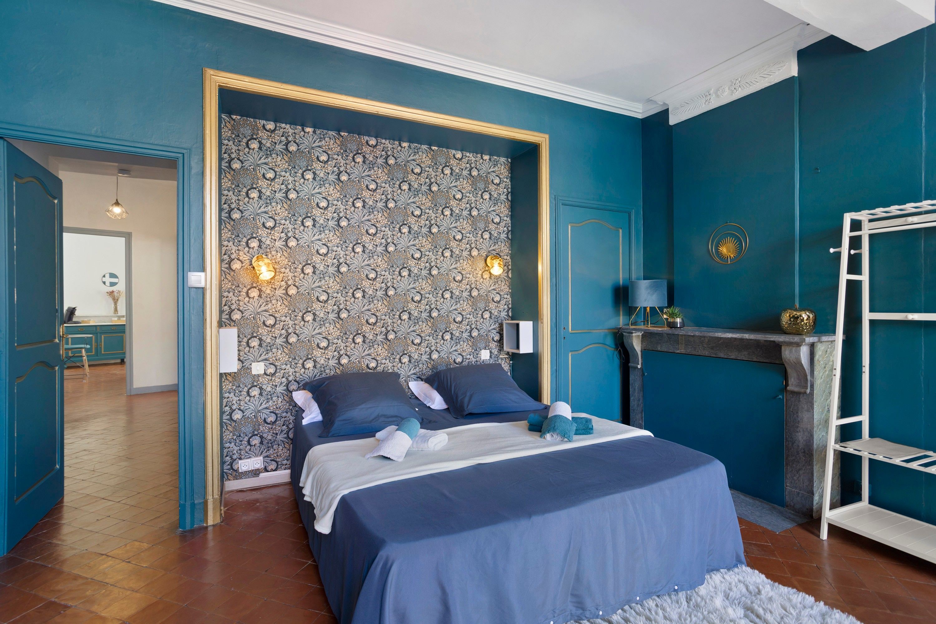 Un dormitorio con papel tapiz estampado, una cama con ropa de cama azul, un radiador blanco y un estante blanco.