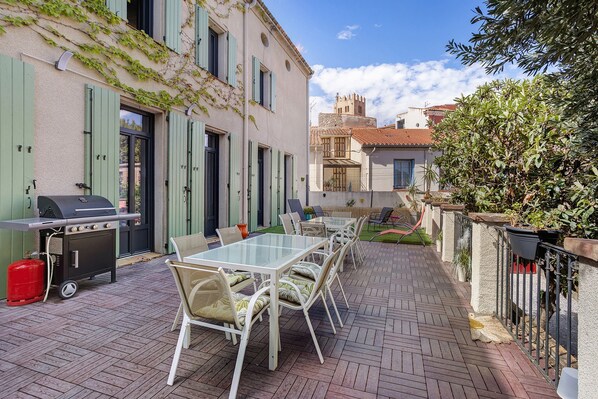 Un patio exterior con una parrilla, mesa de comedor y sillas, rodeado de edificios y follaje.