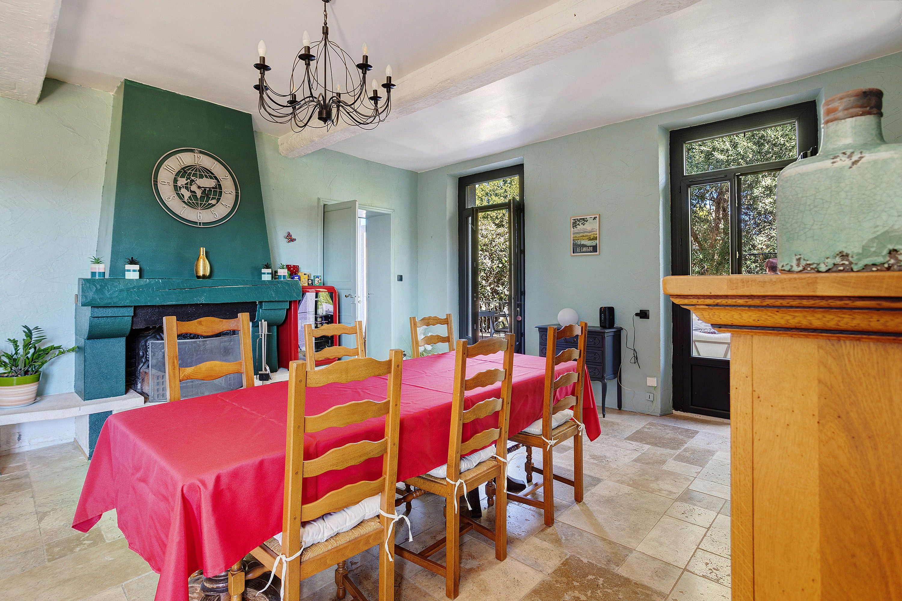 Un comedor con mantel rojo, sillas de madera, una araña y un reloj en la pared.