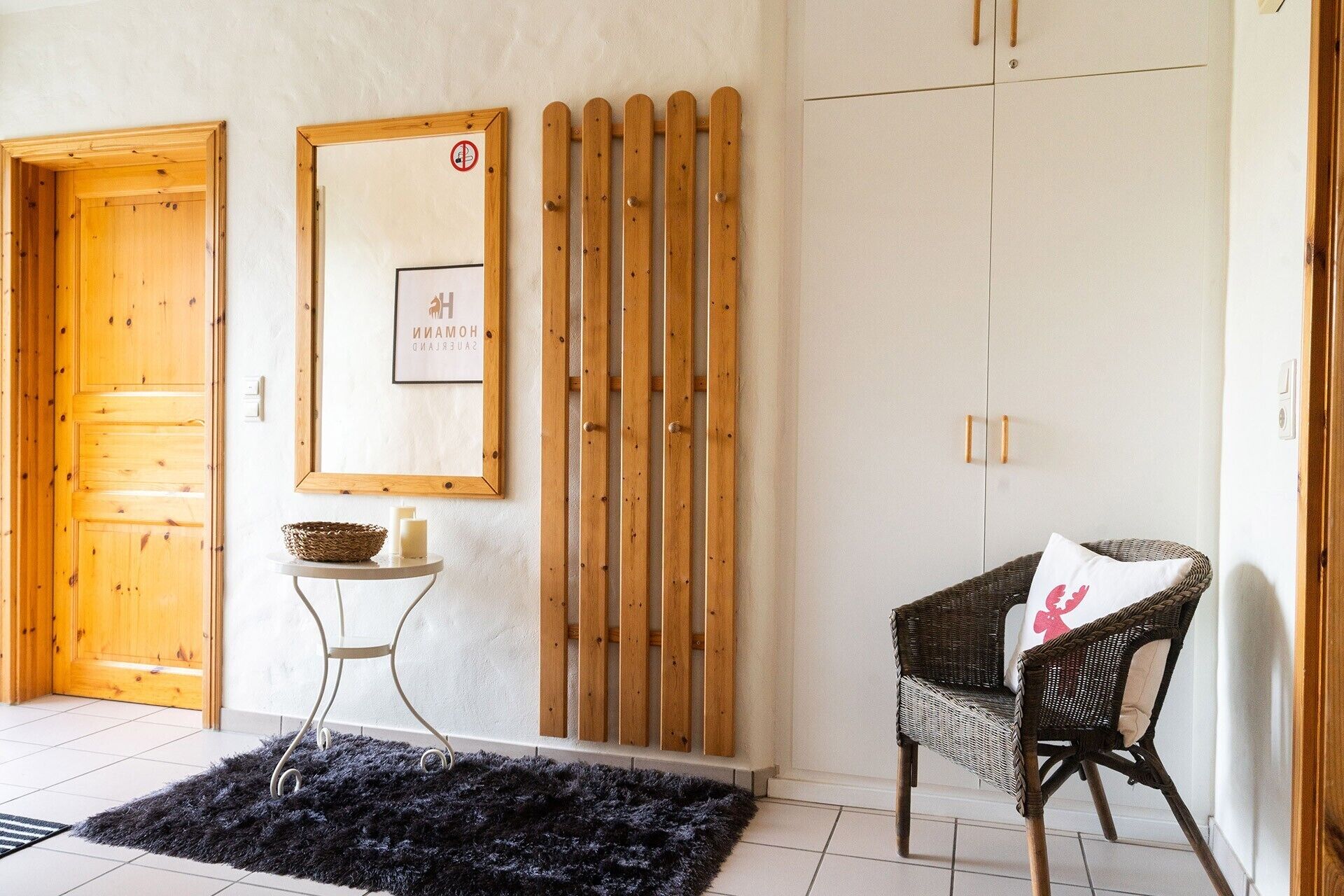 Una habitación con puerta de madera, un cuadro enmarcado en la pared, una silla de mimbre con cojín y un felpudo negro en el suelo.