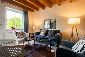 A cozy living room with a sofa, two side tables, a chair, a window with curtains, a radiator, and a framed picture on the wall. - Ferienhaus 'Ferienhof Homann Sauerland – LH 8' mit Gemeinschaftsterrasse und WLAN (Brilon)