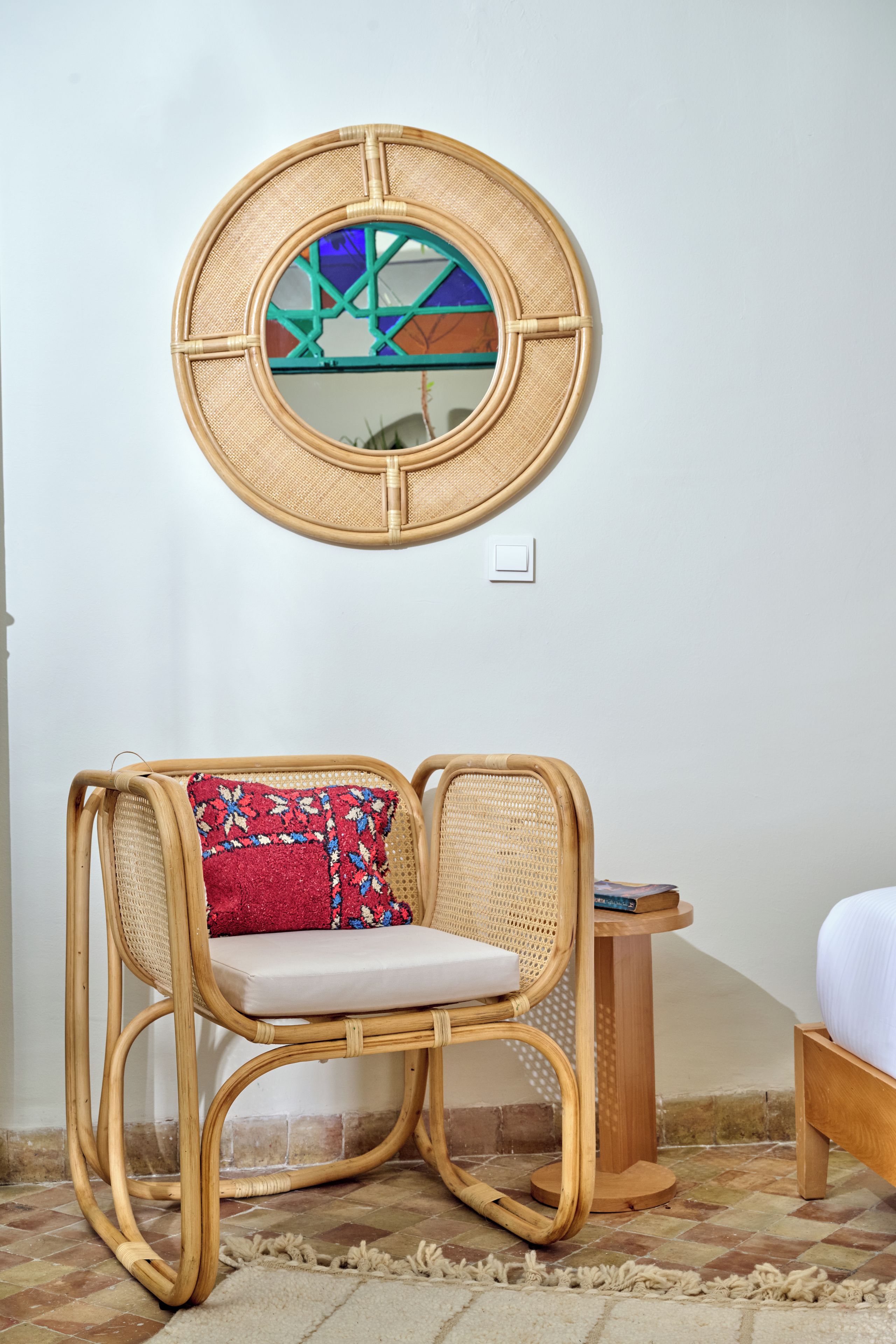 Une chaise en osier avec un coussin, un miroir rond en fibres tressées accroché au mur et une petite table en bois sur laquelle repose un livre.