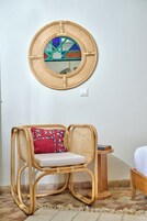 Una sedia in vimini con un cuscino, uno specchio rotondo intrecciato appeso al muro e un piccolo tavolino con un libro.