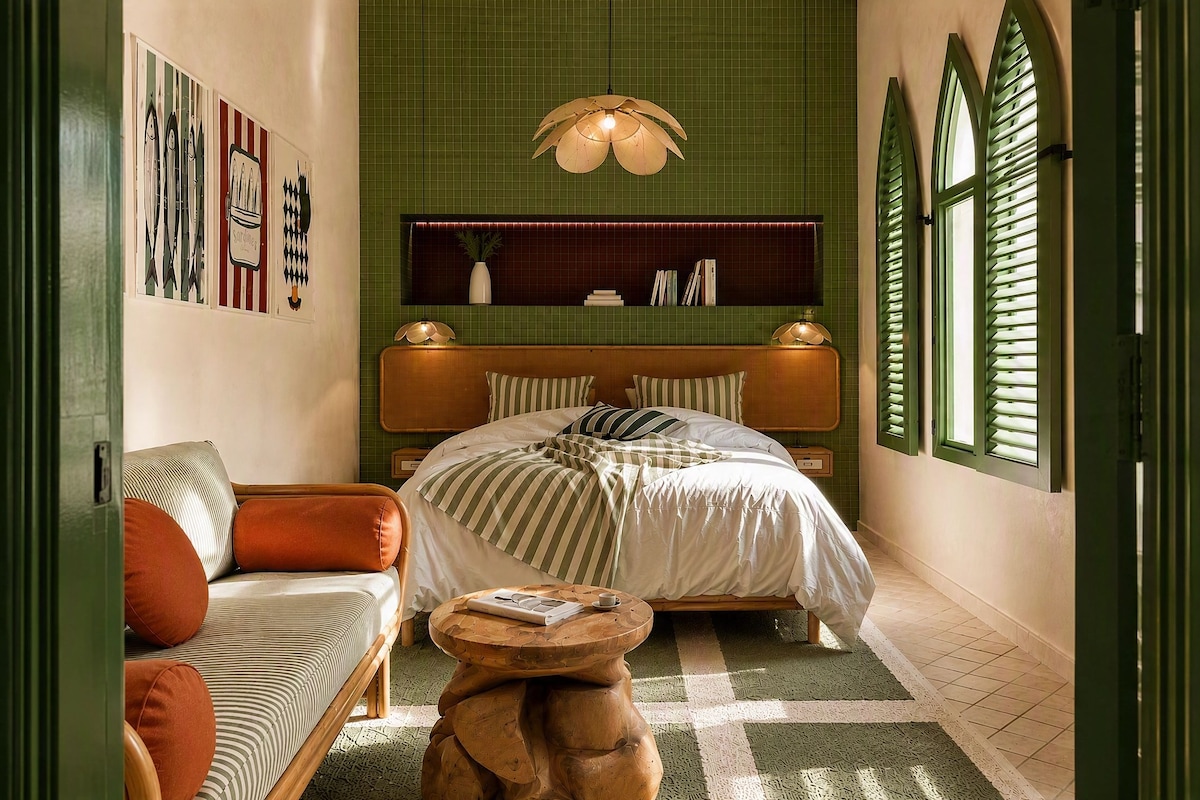 Quarto com cama, sofá, mesa redonda pequena e janela com persianas.