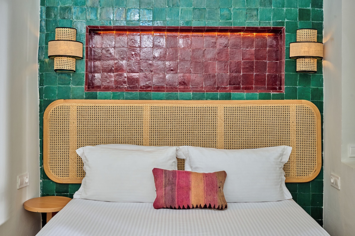 A bed with white linens and a single decorative pillow, a woven headboard, and two wall-mounted lamps.