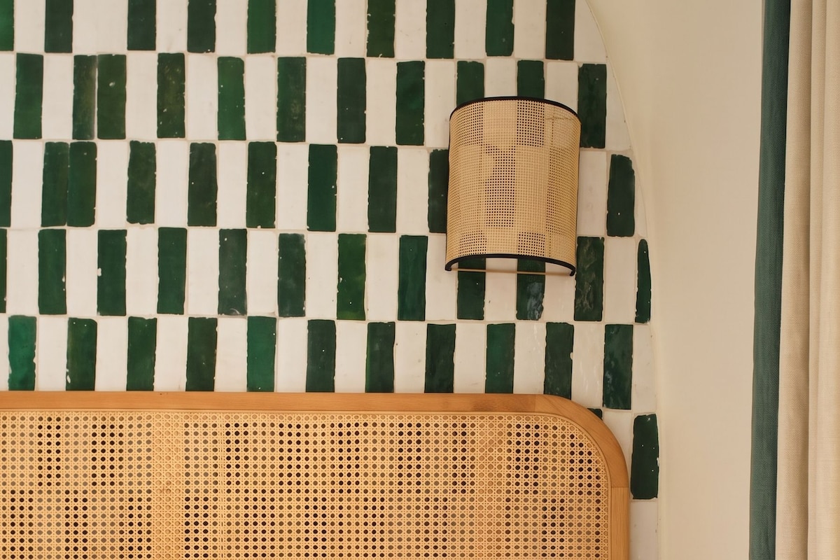 A bed with white linens and a green-striped pillow, a woven headboard, a bedside lamp, a small table with a book and a vase, and a checkered wall.