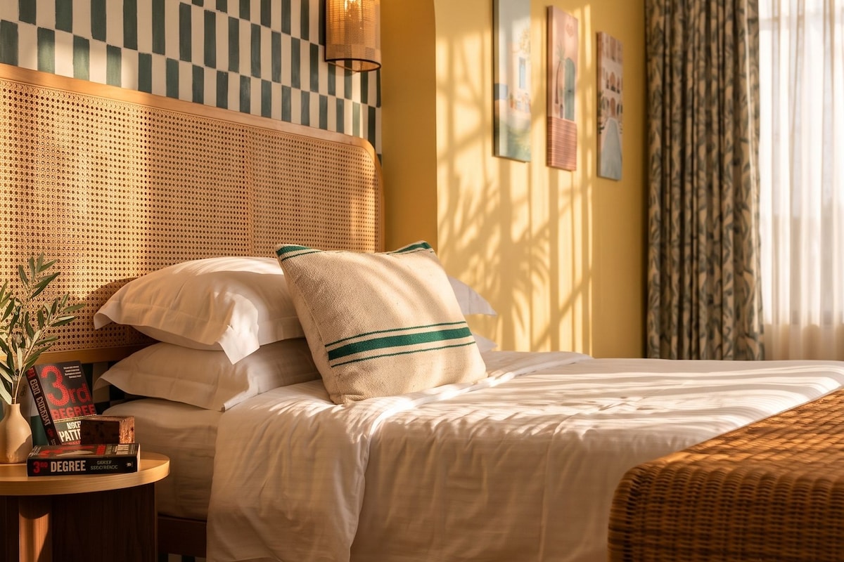 Quarto de hotel com parede xadrez, cama com roupa de cama branca, cabeceira trançada, mesa de cabeceira com livros e uma grande janela com cortinas.
