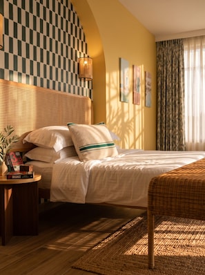 A hotel room with a checkered wall, a bed with white linens, a woven headboard, a bedside table with books, and a large window with curtains. - StayHere Essaouira – Riad Heritage (Essaouira)