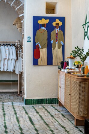 A hotel lobby with a wall-mounted painting, a rack of white shirts, a wooden cabinet with food items, and a patterned floor.