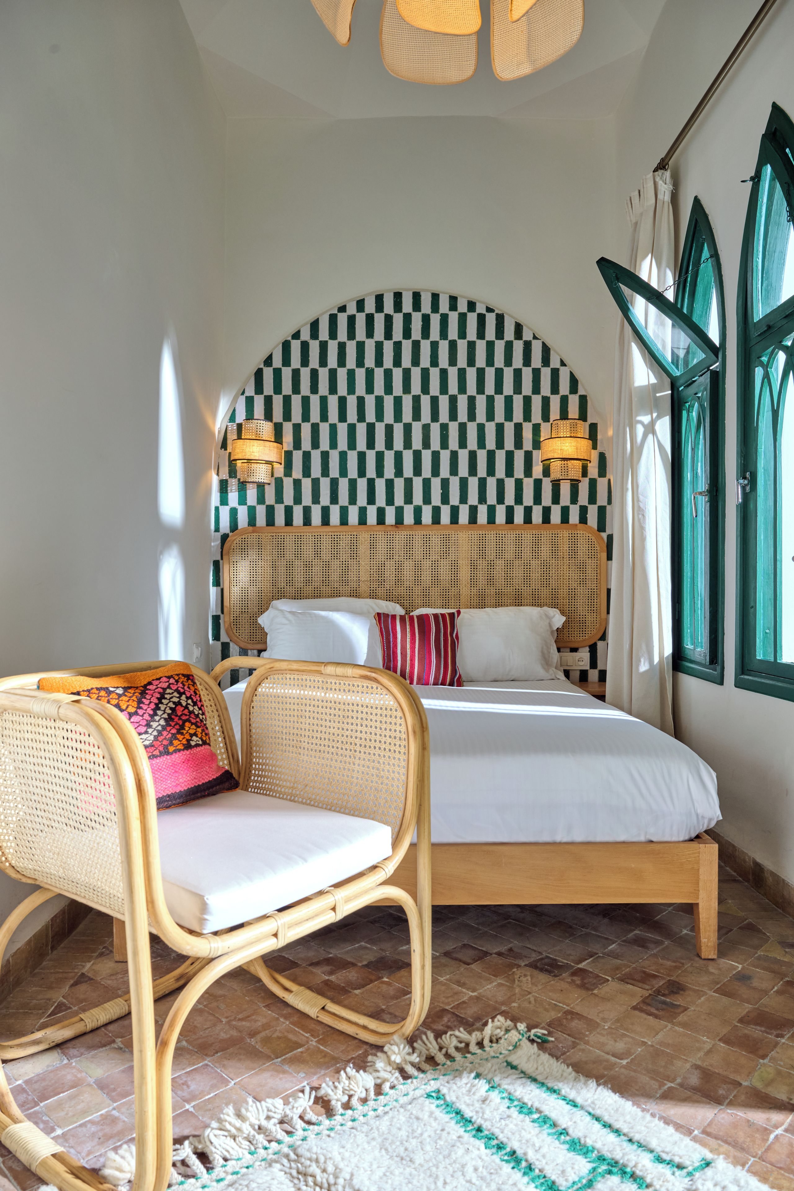 Une chambre avec un mur à carreaux, un lit recouvert de draps blancs, une chaise en osier et une fenêtre avec des volets verts.