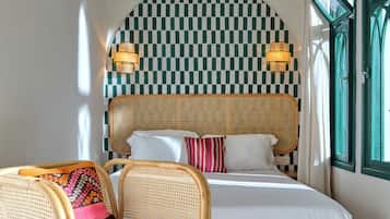 Une chambre avec un mur à carreaux, un lit recouvert de draps blancs, une chaise en osier et une fenêtre avec des volets verts.