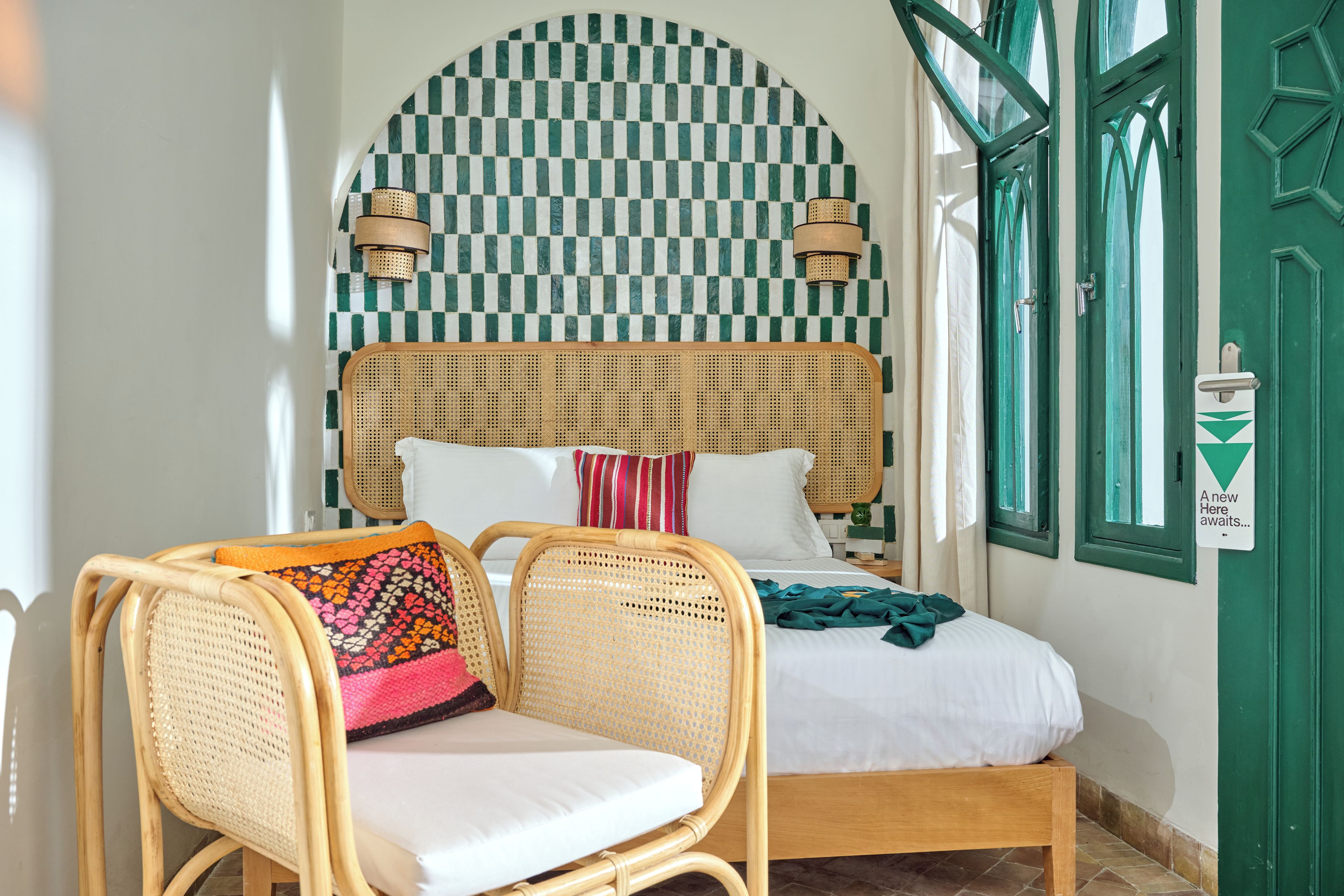 Una habitación con pared a cuadros, una cama con ropa de cama blanca, una silla de mimbre con un cojín colorido y una puerta verde con un cartel.