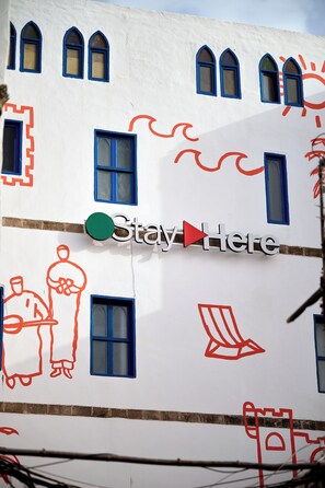 A building facade with multiple windows, some with blue frames, and a large sign with the text 'Stay Here' and a green circle. - StayHere Essaouira – Riad Heritage (Essaouira)