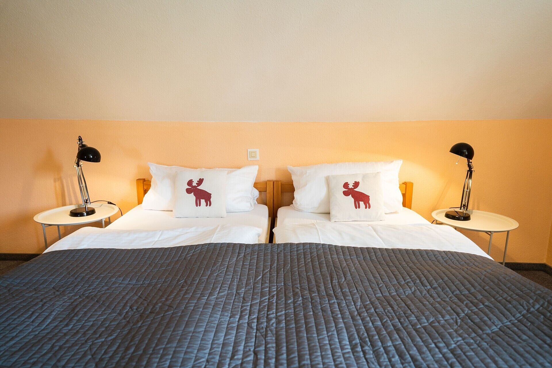 A double bed with a grey quilted cover, two white pillows with a moose design, and two bedside lamps on matching nightstands.