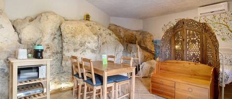 A room with a stone wall, a wooden dining table with chairs, a coffee machine, a microwave, and a wooden cabinet.