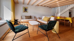 Ett modernt vardagsrum med synliga träbjälkar, en soffa, två fåtöljer, ett soffbord och en matplats med en gul duk.