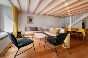 Ein modernes Wohnzimmer mit Holzbalken, einer Couch, zwei Sesseln, einem Couchtisch und einem Essbereich mit gelber Tischdecke.