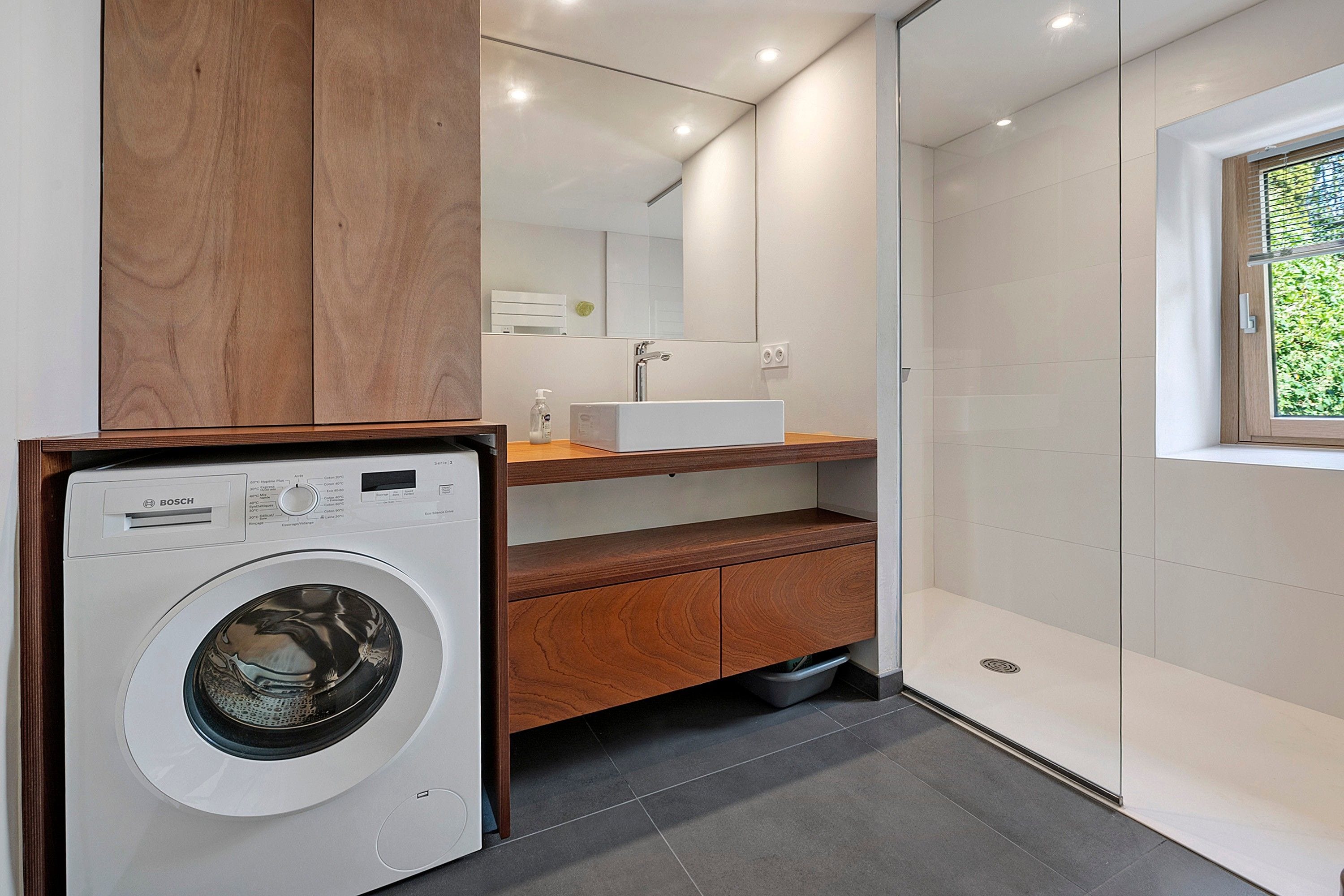 Een moderne badkamer met een Bosch wasmachine, een houten wastafelmeubel, een spiegel en een douchehoek.