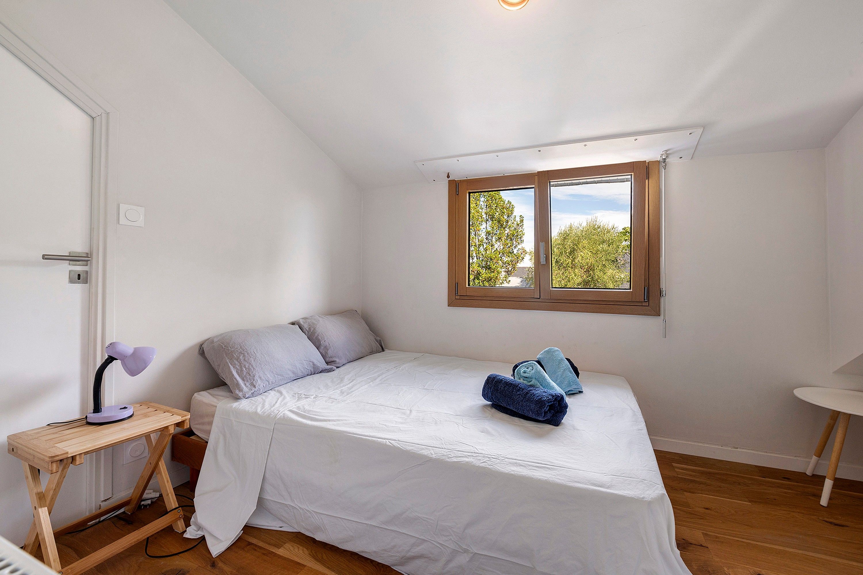 A small, single-bed room with a wooden floor, a desk with a lamp, and a window with a view of trees.