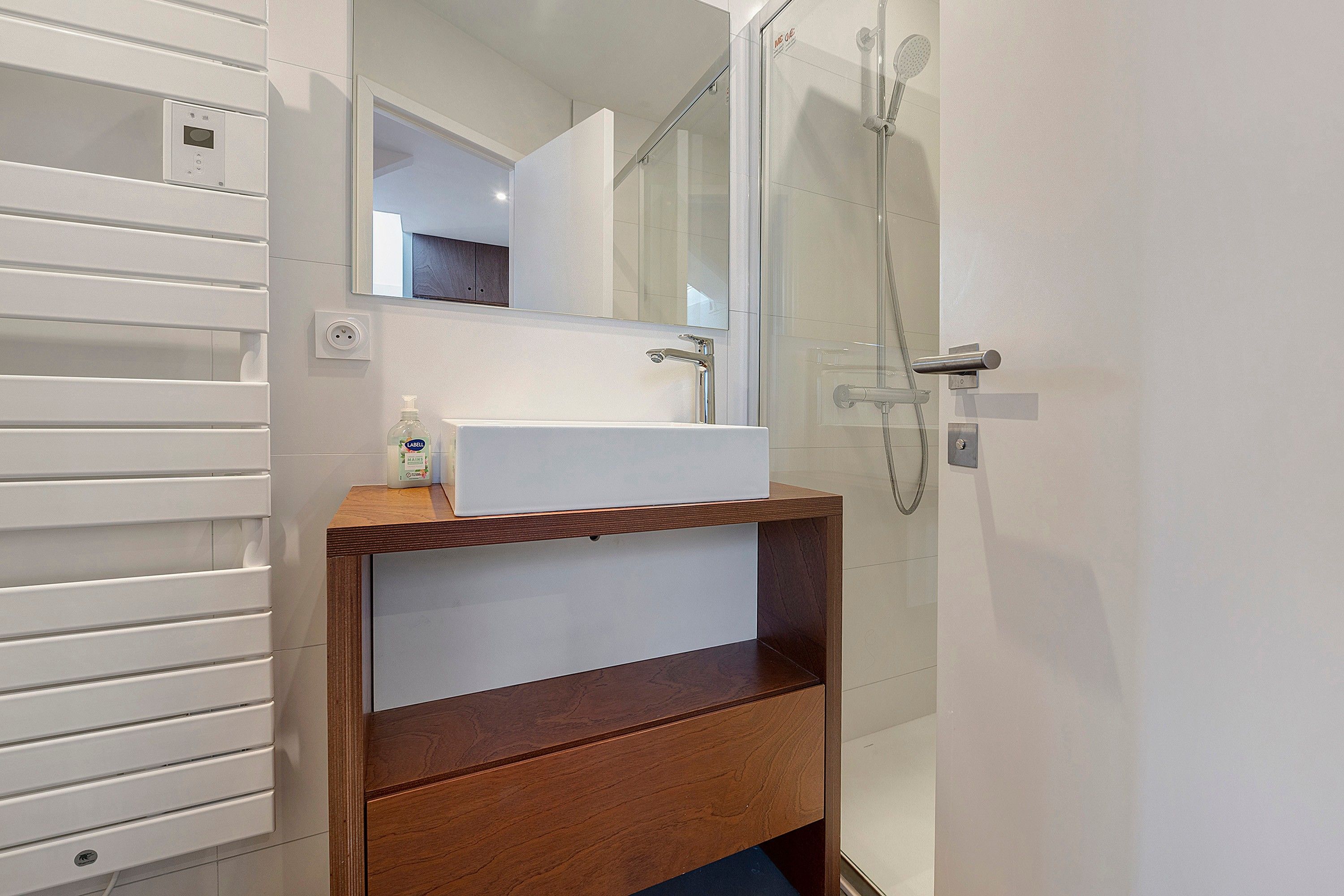 Une salle de bain avec un lavabo blanc, un miroir au-dessus, un espace douche et un meuble-lavabo en bois sur lequel se trouve un flacon de savon liquide.
