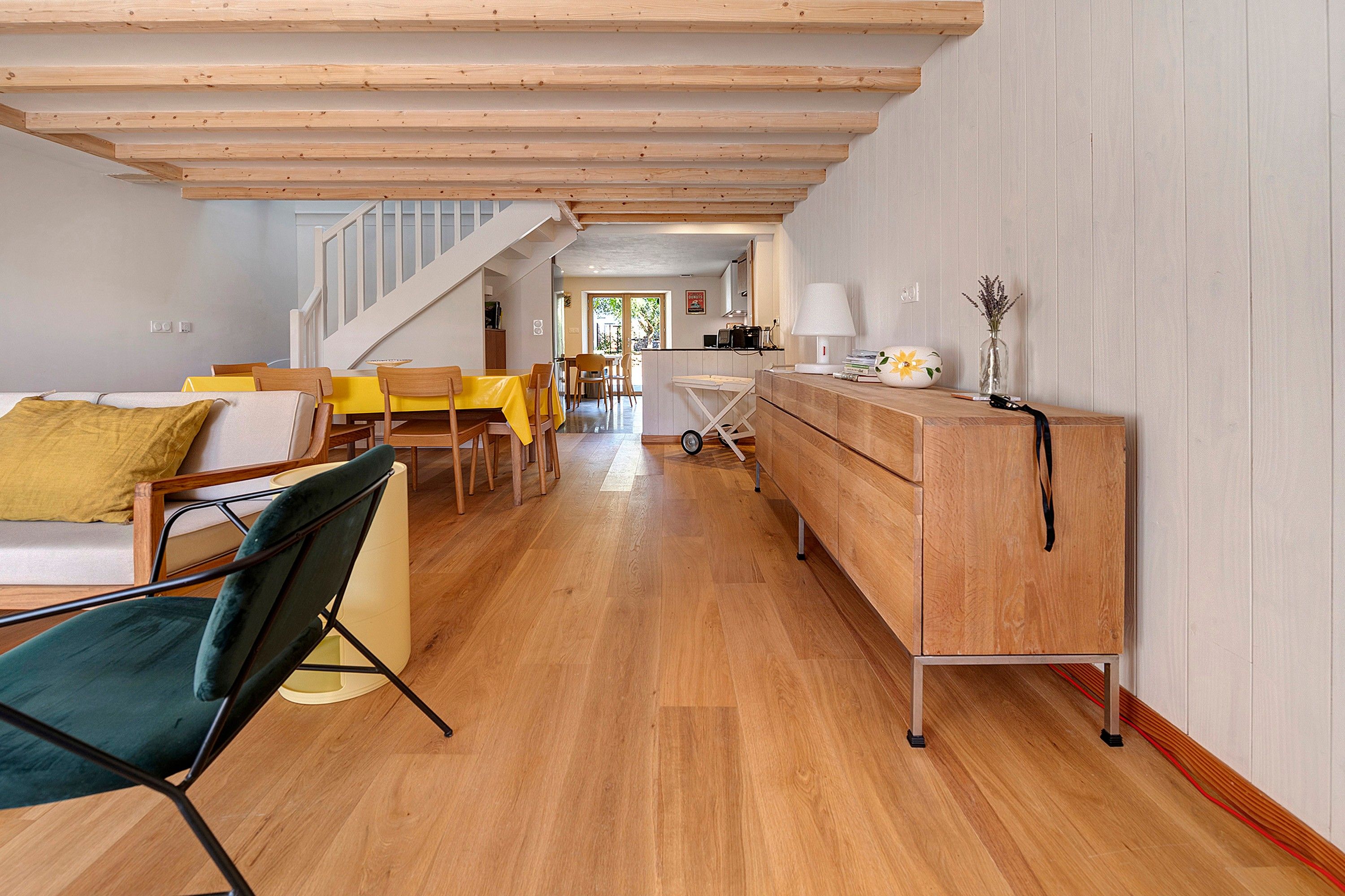 A spacious living area with a wooden floor, a staircase leading to another level, a dining area with yellow tables, and a kitchen visible in the background.
