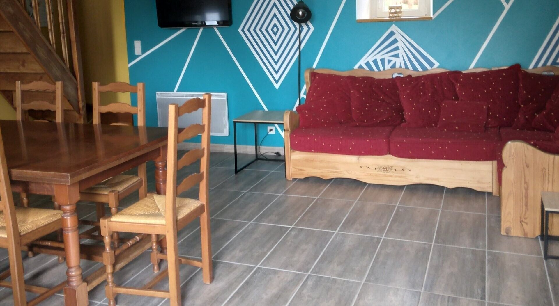 Un comedor con mesa y sillas de madera, un sofá con cojines rojos y una pared con diseños geométricos.